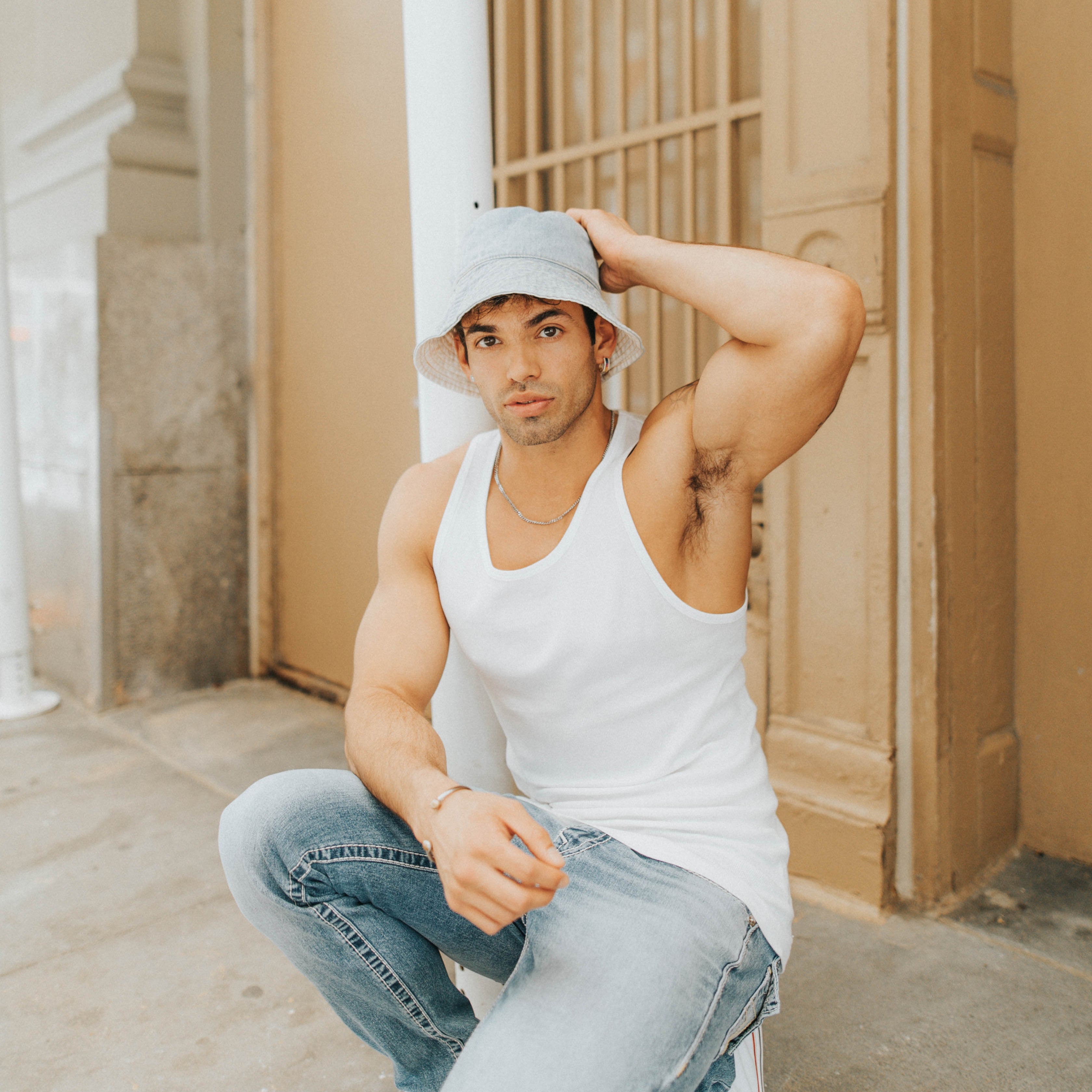Male model crouched on a sidewalk in New York City wearing light blue jeans, a white tank top, and a white bucket hat with one hand on the hat