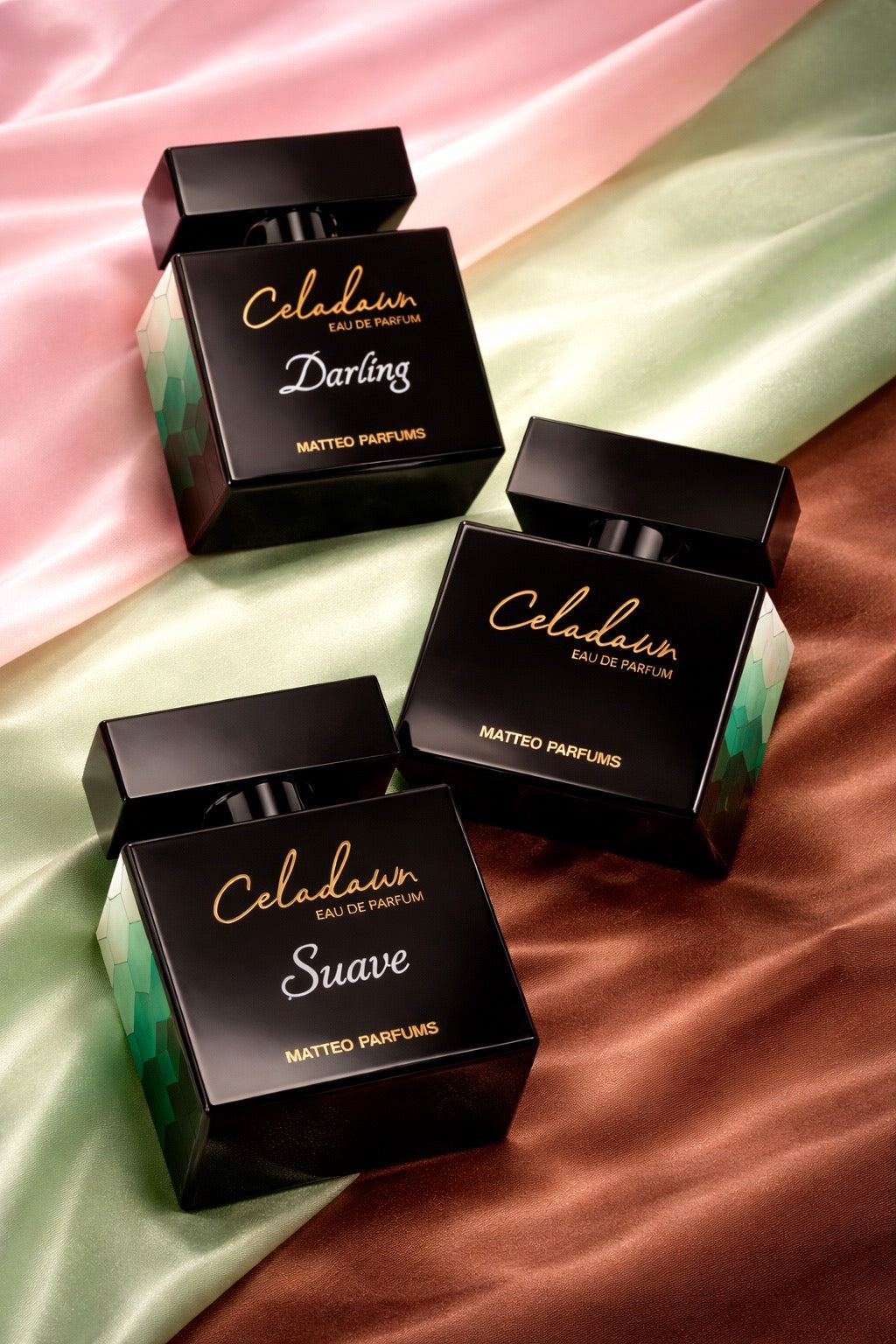 Three black perfume bottles "Celadawn", "Celadawn Darling", and "Celadawn Suave", written in gold text laying on a multi-colored pink, green, and brown satin surface
